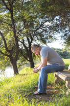 a man sitting with his head bowed in prayer on a bench by a lake 