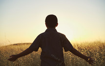 a young man with outstretched arms walking through a field of tall grass at sunset 