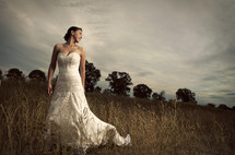 bride standing in a field - tree background - cloudy sky