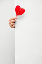 Male hand holding and offering Heart Shaped Lollipop behind white wall background