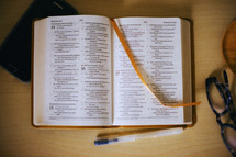 A bible, glasses,  phone, and pen laid out for reading. 