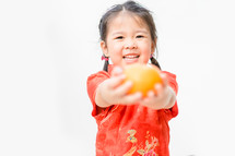 A little girl in a traditional Chinese dress holding out an orange 