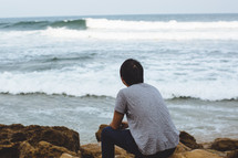A man sitting on a rocky shore looking out at the ocean.