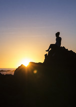 a silhouette of a woman sitting on a rock on a shore at sunset 