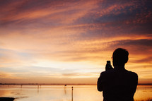 Silhouette of a man taking a picture of a sunset with his cell phone.