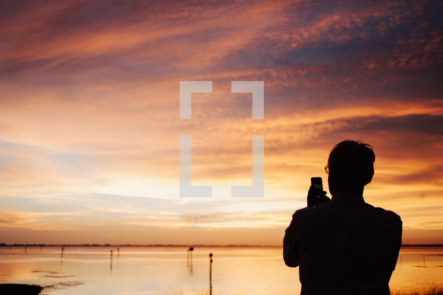 Silhouette of a man taking a picture of a sunset with his cell phone.