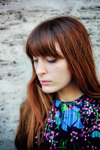 Young woman with long brown hair and freckles, wearing a floral print top, is looking down with a thoughtful expression