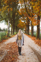 woman walking down a rural dirt road covered in fall leaves 