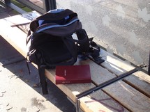 bible next to a backpack on a bench at a bus stop, 

bible, bench, stop, backpack, bus stop, waiting, tram, station, streetcar, trolley, transportation, way, bus, bag, rucksack, knapsack, booksack, daybag