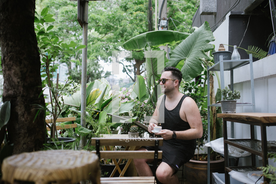 a man sitting at an outdoor table surrounded by tropical foliage drinking an espresso