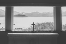 A cross in a window with a view of a lake and mountains.