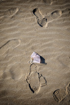 Surgical mask is laying on the sand with footprints surrounding it, suggesting it was discarded