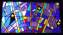 A colorful stained glass window with blue, lavender, purple, red and gold and symbols adorn a church worship center in a prayer chapel. 