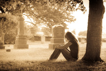 woman sitting in front of a tree in a cemetery grieving 