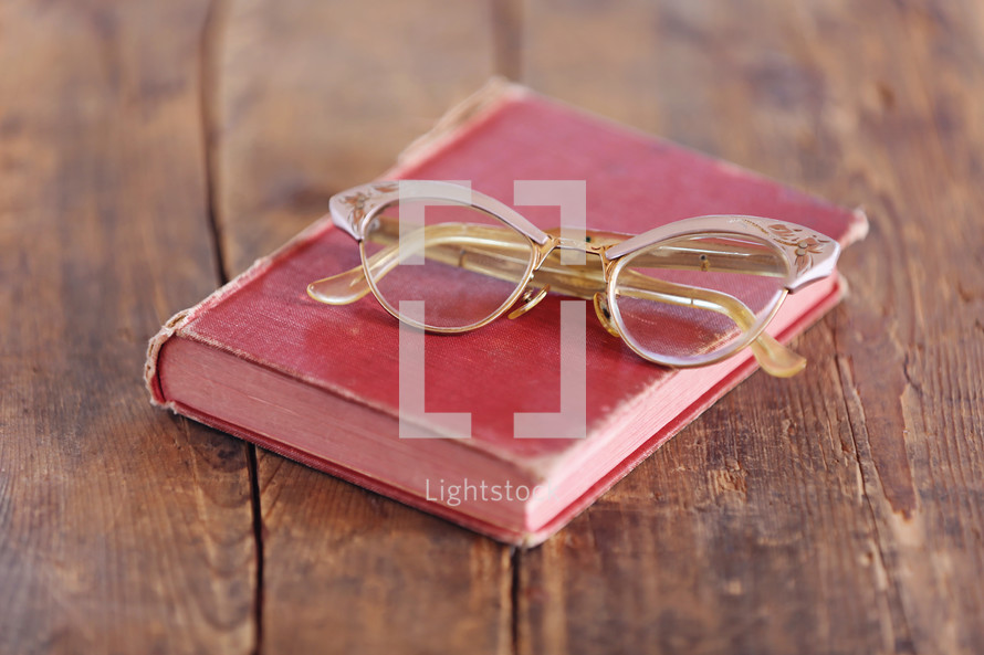 Eye glasses on an old red book which is on an rugged wooden table.