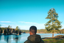 a man in a coat standing with his back to the camera looking out at a lake 