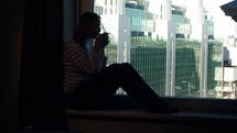 Woman drinking coffee on the windowsill with city view in window