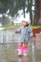 Toddler girl wearing coat, hat and rainboots in rain on wet sidewalk.