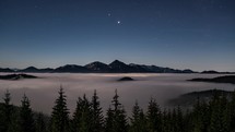 Blue nigh sky with stars above alpine mountains, forest and foggy clouds motion fast in moonlight evening nature landscape Time lapse
