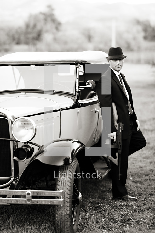 gangster man in front of an old car holding a tommy gun