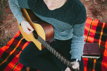 man with a guitar sitting on a plaid blanket 