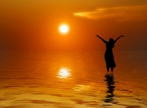 Silhouette of woman standing in water praising God at sunrise.