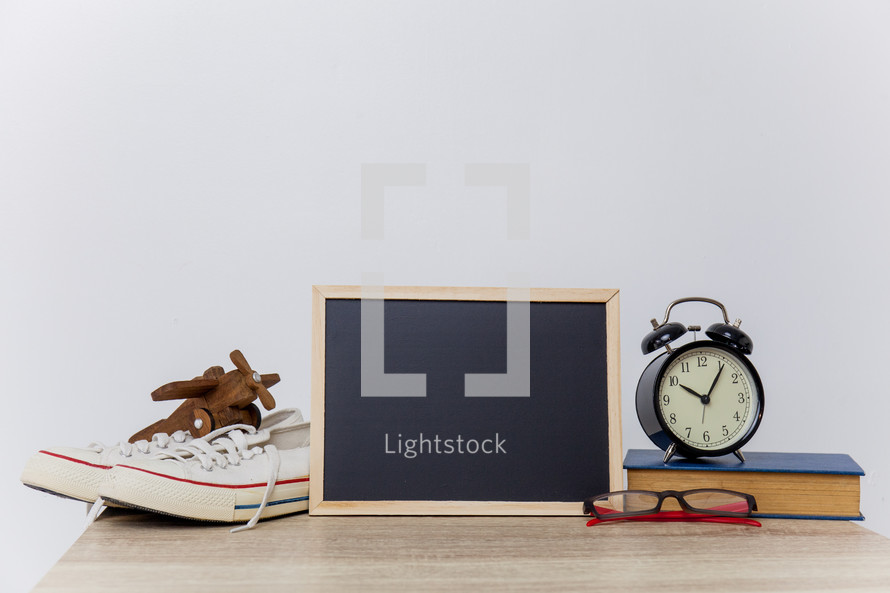 white sneakers, chalkboard, wooden toy plane, alarm clock, book, glasses, table 