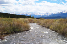 A river flowing through a field toward snow capped mountains with blue sky and clouds