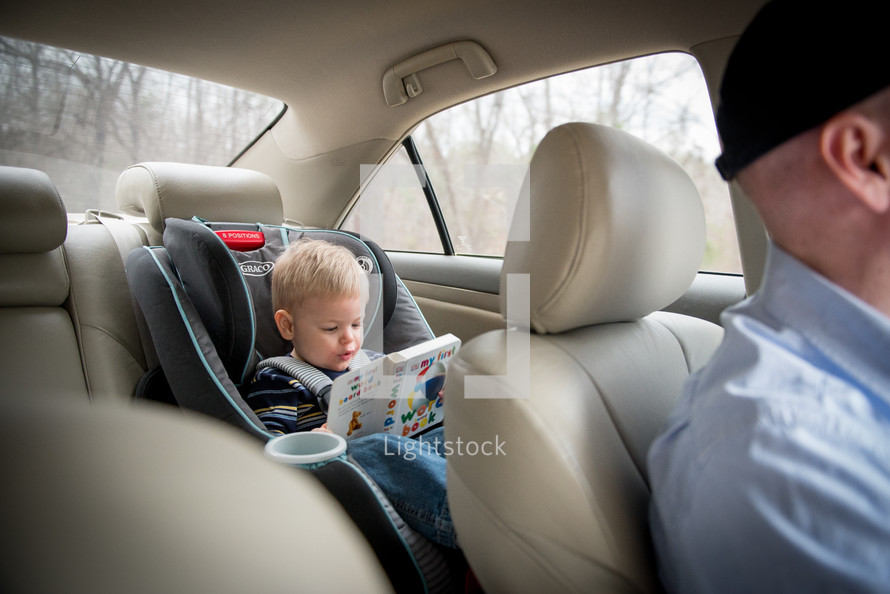 toddler boy reading a book in a carseat 