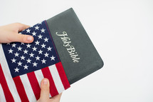 Hands holding a Holy Bible wrapped in an American flag.