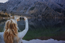 a woman taking a picture of a lake with her phone 