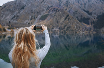 a woman taking a picture of a lake with her cellphone 