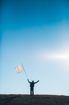Silhouette of man waving white flag with arms raised on grassy hilltop.