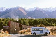 A white van parked in front of a log house at the base of a mountain range.