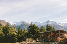A shed full of firewood surrounded by trees and a mountain range.