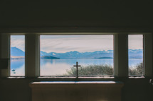 A cross in a window looking out at a lake and mountains.