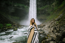 a woman wrapped in a blanket standing in front of a waterfall 