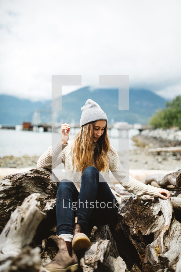 a woman in a wool cap sitting on driftwood on a beach 