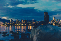 woman sitting on a rock and a view of a coastal city skyline at night 