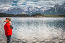boy standing on a lake shore with mountains in the background 