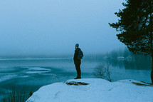 man standing on a snowy rock on a lake shore 
