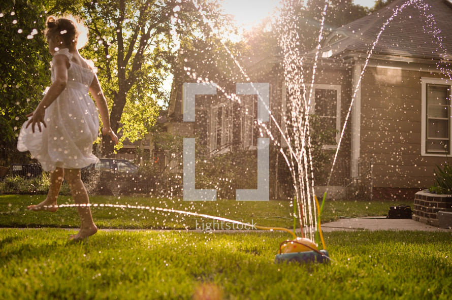 A little girl dances in the sprinklers in the warm golden light of summer.