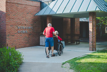 husband pushing his wife in a wheelchair in front of a nursing home