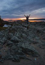a man with raised hands standing on a rocky shore at sunset 