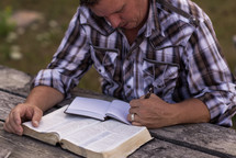 a man reading a Bible and writing in a journal at a picnic table outdoors 