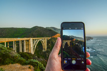 a man taking a picture with a cellphone of a bridge over a ravine along a shoreline 