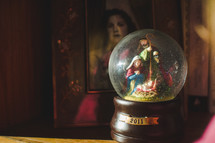 A Christmas nativity snow globe sitting on a display case with a framed family photo behind it.