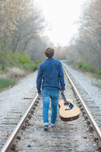 Man walking on a railroad track holding an acoustical guitar.