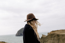 A blonde woman in a hat stands near the ocean.