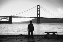 Silhouette of a man looking at a suspension bridge.